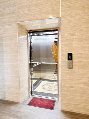 Een lift met een metalen interieur, een rode mat bij de ingang en een bedieningspaneel aan de muur.
