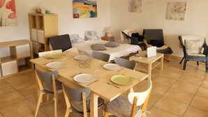 A dining area with a wooden table set for four, surrounded by chairs. There is a bed with floral bedding in the background, a lamp on the nightstand, and artwork on the walls.
