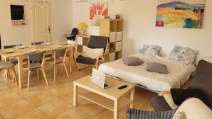 A modern, well-lit room with a dining area, a bed with multiple pillows, a laptop on a table, and a painting on the wall.
