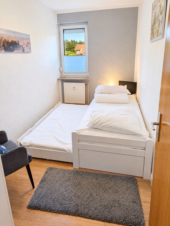 A small, single-bed room with a window, a desk, and a grey rug.
