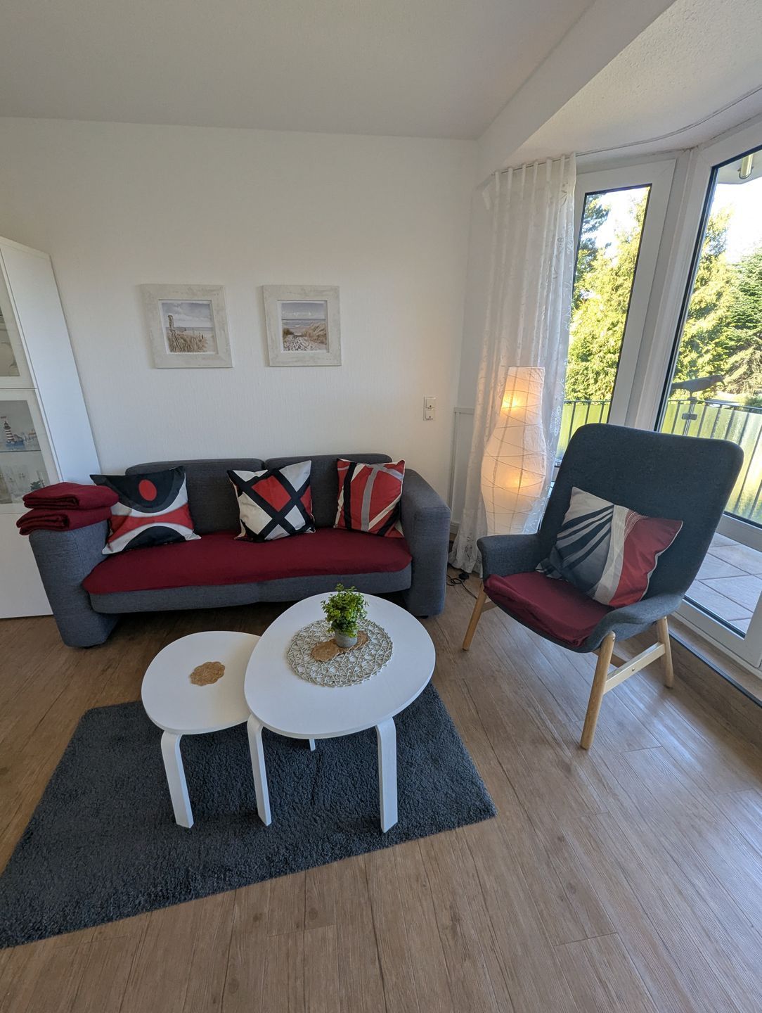Ein Wohnzimmer mit grauem Sofa, einer roten und schwarzen Decke mit Muster, einem kleinen weißen Tisch mit Pflanze und einem hölzernen Stuhl mit rotem Kissen.