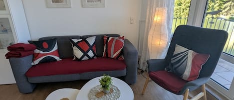 A living room with a grey sofa, a red and black patterned throw, a small white table with a plant, and a wooden chair with a red cushion.