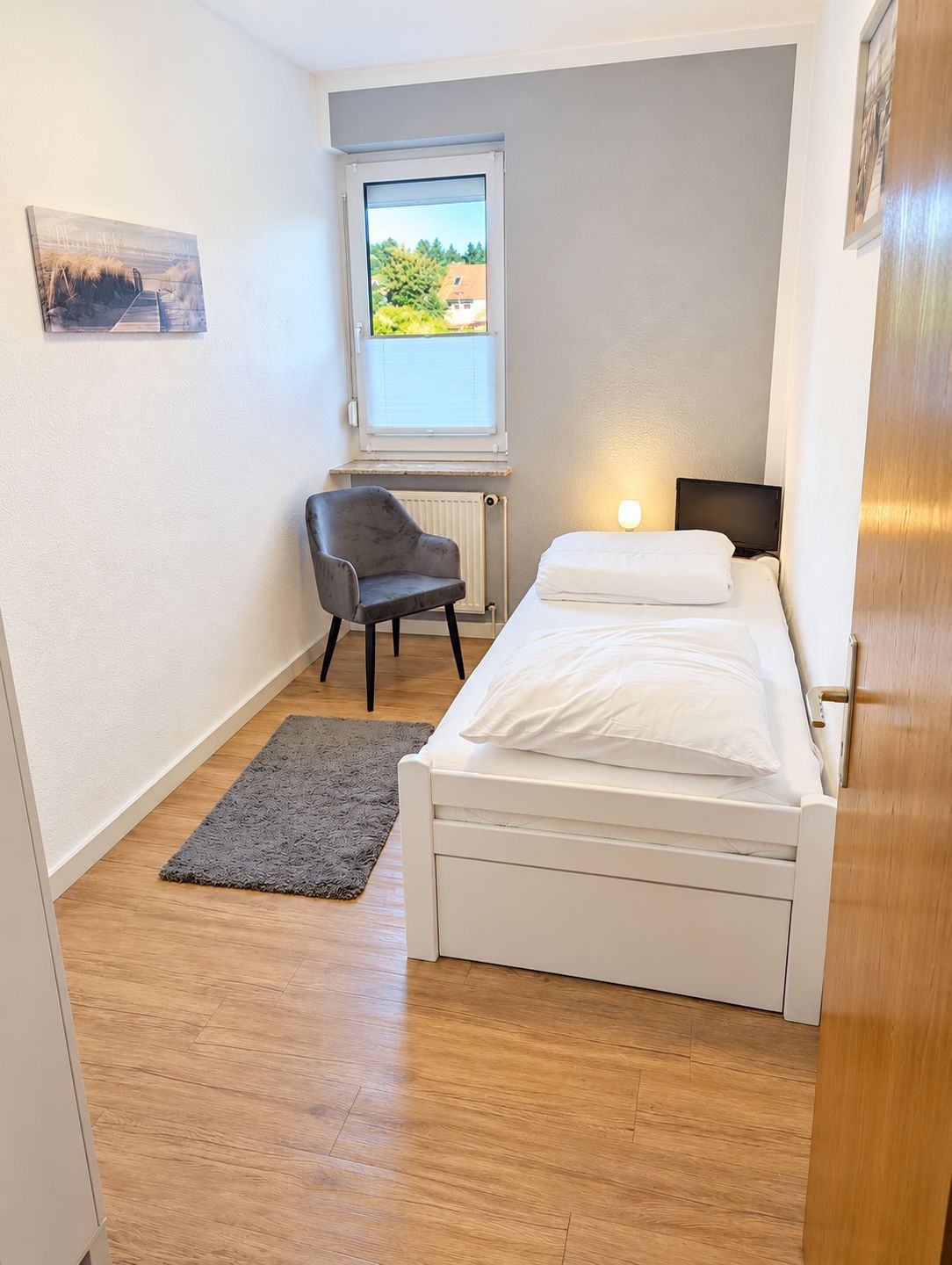 Ein kleines, ordentliches Schlafzimmer mit einem Einzelbett, einem Sessel, einem Fenster mit Blick und einer Heizung.