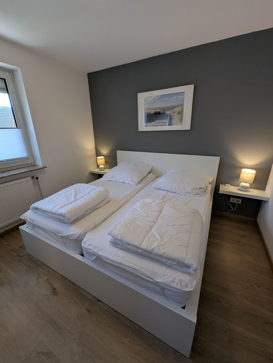 Ein Doppelbett mit weißer Bettwäsche, zwei Nachttische mit Lampen, ein gerahmtes Bild an der Wand und ein Fenster mit Jalousien.