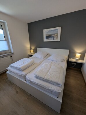 Una cama doble con ropa de cama blanca, dos mesitas de noche con lámparas, un cuadro en la pared y una ventana con persianas.