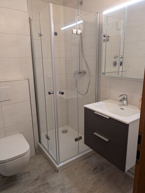 Un baño moderno con ducha de vidrio, inodoro blanco, lavabo con espejo y mueble oscuro.