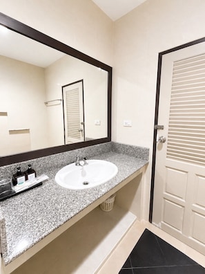 A bathroom with a white sink, a large mirror, and a door with horizontal blinds. - Relax Corner Jomtien – Spacious Studio with Balcony (Muang Pattaya)