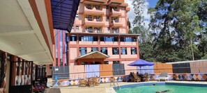 A multi-story hotel with a pool area, outdoor seating, and a view of trees. - Pennjoki Resort (Kakamega)