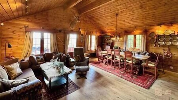 A cozy cabin interior with wooden walls, a dining area, and a living room with a sofa and chairs.