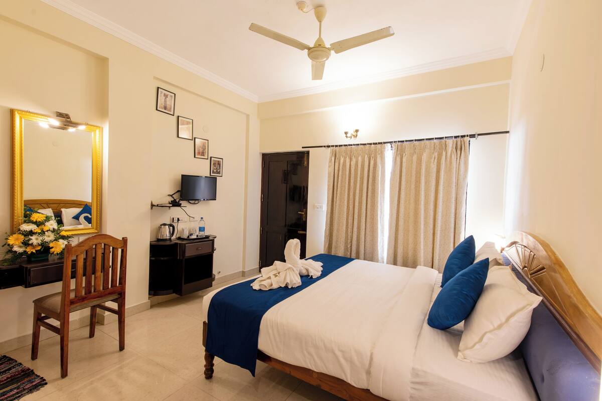 A hotel room with a bed, a wooden chair, a desk with a lamp, a television, and a ceiling fan.