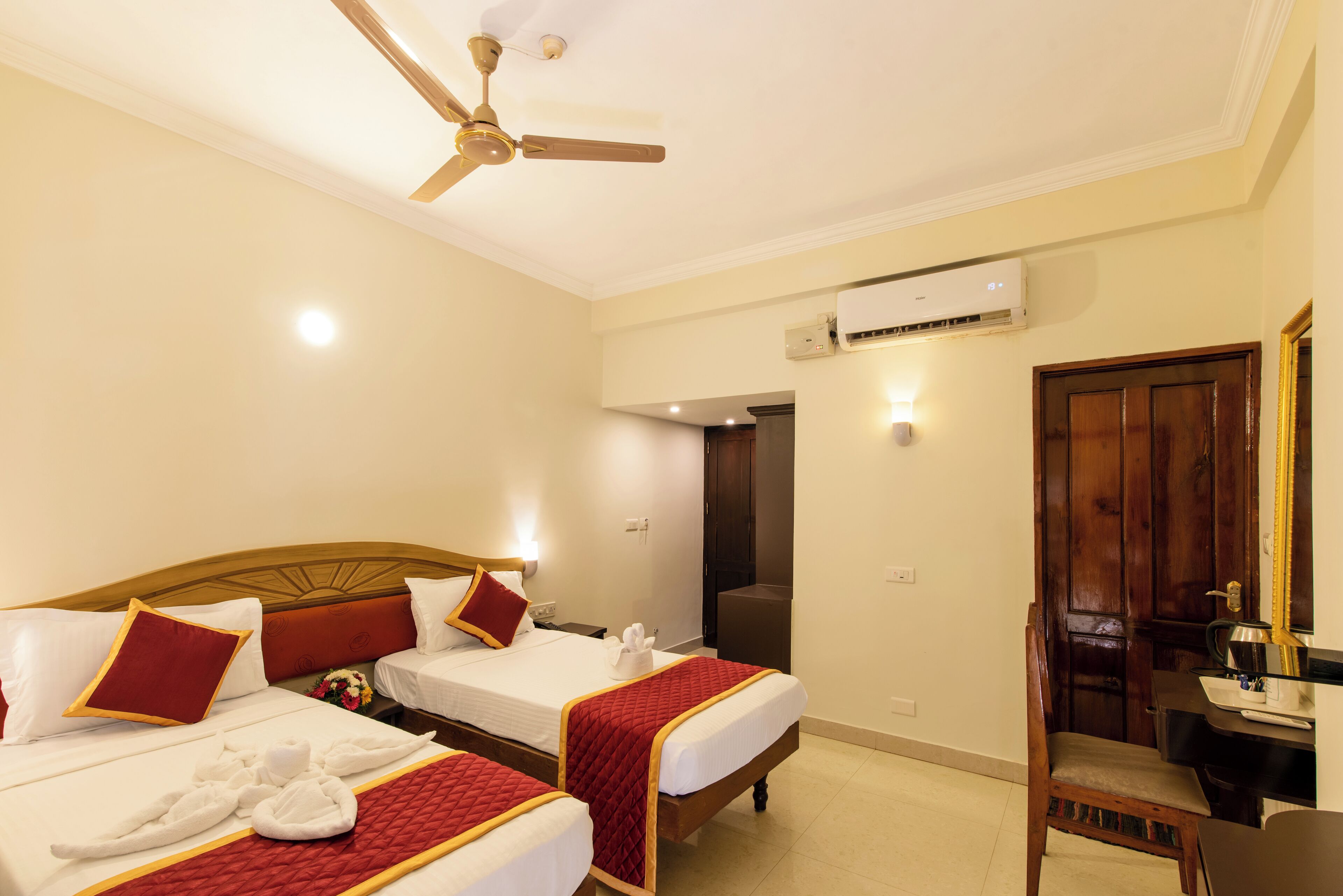 Habitación de hotel con dos camas, ventilador de techo, aire acondicionado, escritorio con silla y puerta de madera.