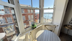 A room with a large bay window overlooking a cityscape and water. There is a round table with a chair, a small plant on the windowsill, and a lamp on a side table. - Merchants Quarters - Laguz & Othala - sleeps 8 guests  in 4 bedrooms (Scarborough)