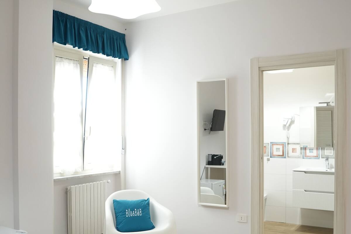 A modern hotel room with a bed, a chair with a blue pillow, a white radiator, and a view into the bathroom.