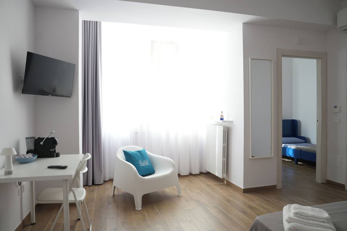A modern hotel room with a flat-screen TV, a desk with a coffee maker, a white armchair with a blue pillow, a large window with curtains, a radiator, and a blue chair in the background.