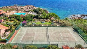 A tennis court surrounded by lush greenery and a pool, with a view of the sea and rocky coastline. - Beautiful apartment for 6 people with pool, WIFI, A/C, TV and patio (La Maddalena)