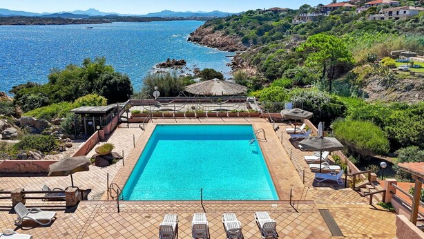 A rectangular swimming pool with a tiled deck, surrounded by lounge chairs and umbrellas, overlooking a coastal area with hills and a clear blue sea. - Beautiful apartment for 6 people with pool, WIFI, A/C, TV and patio (La Maddalena)