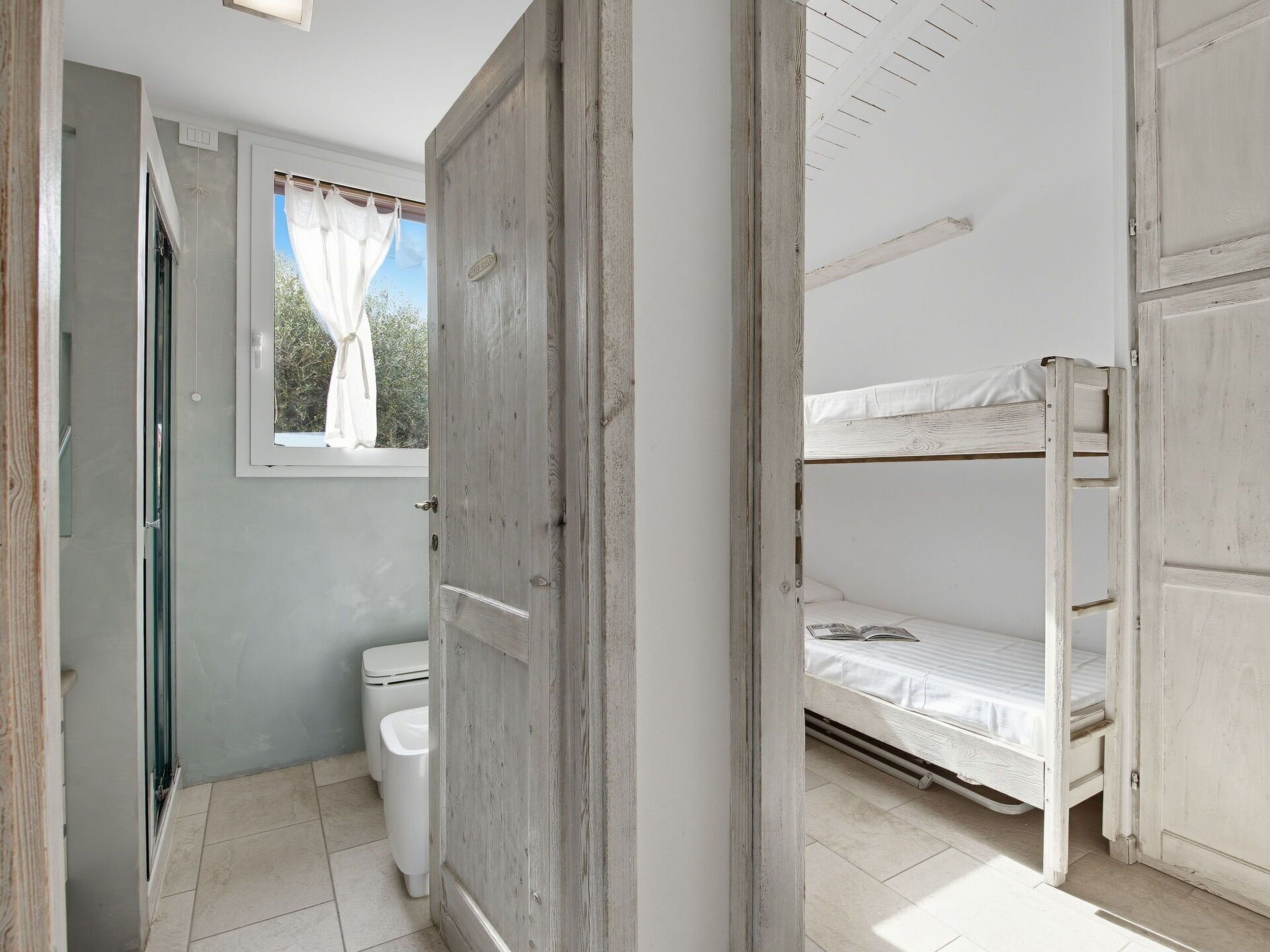 Een kleine, rustieke badkamer met een houten deur, een raam met gordijnen en een wit toilet.