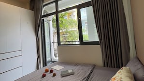 A bedroom with a large window, a bed with a grey mattress, a bedside table with a book and fruit, and a wardrobe. - Unite StayCation Le Van Sy (Ho Chi Minh City)