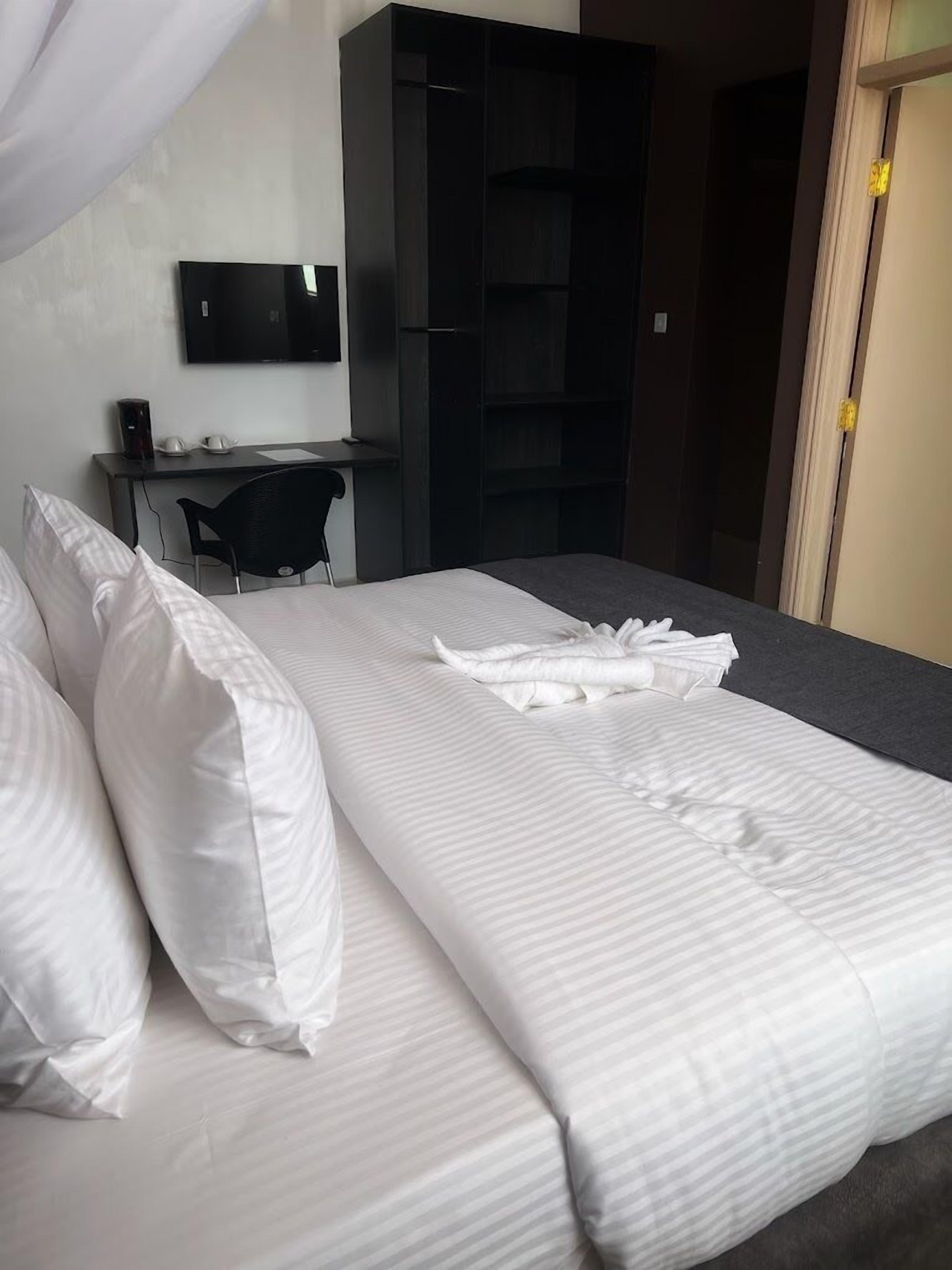 A neatly made bed with white linens, a desk with a computer and a chair, and a TV mounted on the wall.