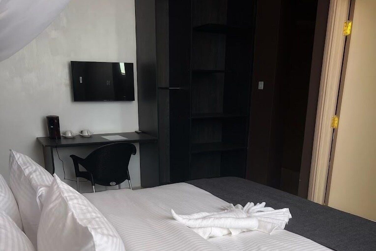 A neatly made bed with white linens, a desk with a computer and a chair, and a TV mounted on the wall.