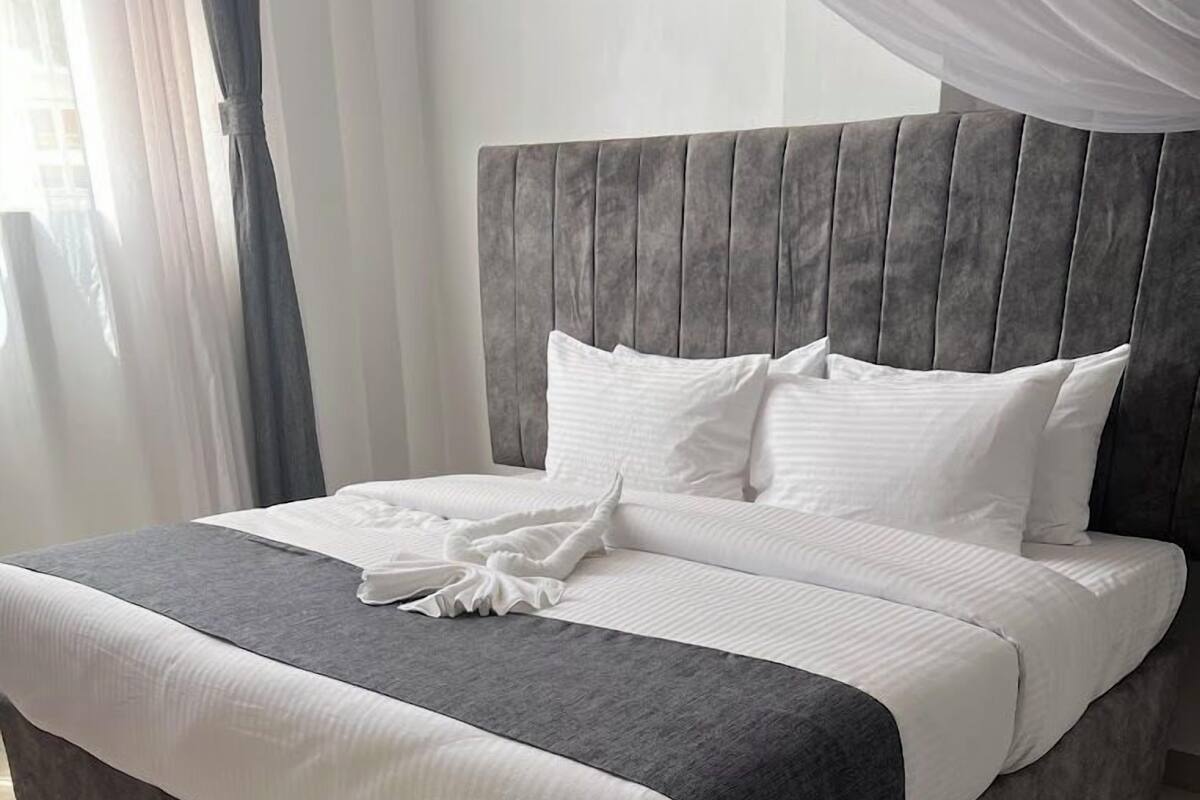 Una cama bien hecha con un cabecero tapizado en gris, ropa de cama blanca y una manta gris.