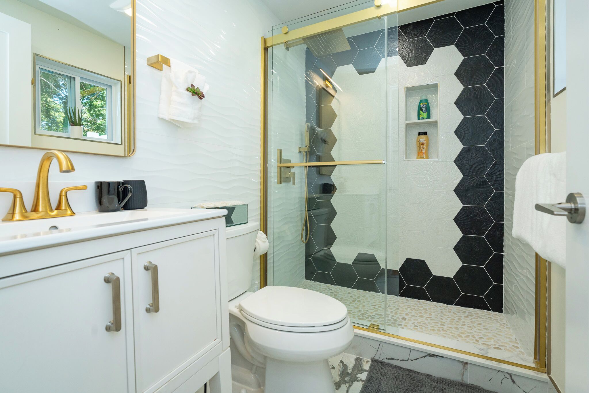 A modern bathroom with a white toilet, a gold faucet, and a glass-enclosed shower with black hexagonal tiles.
