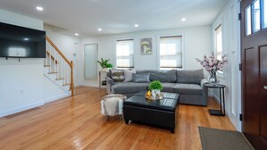 A spacious living room with hardwood flooring, a grey sofa, a black coffee table, and a television mounted on the wall. - Elegant 4BR/3BR Home w/Yard & 4 Parking in Dedham (Dedham)