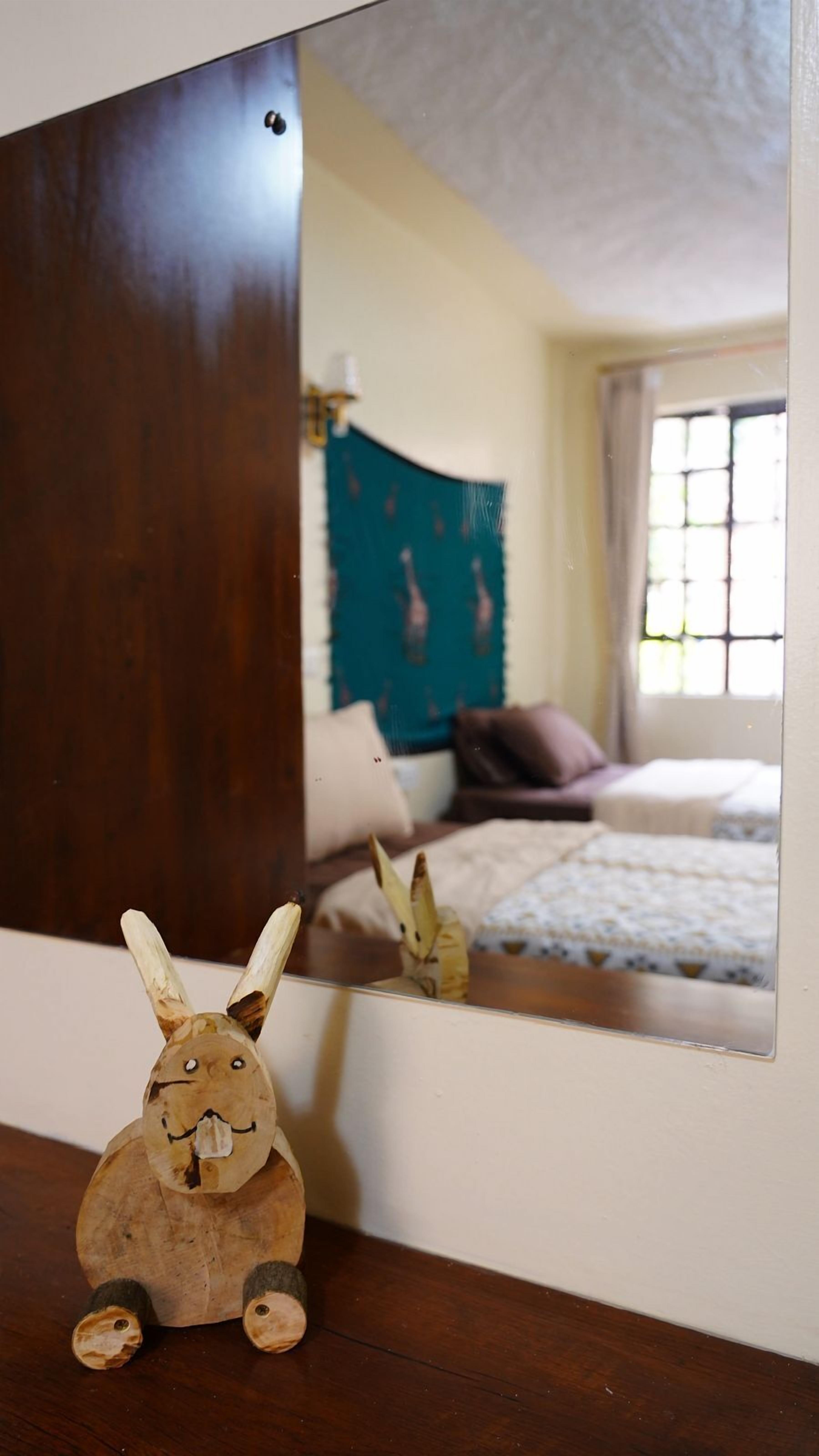 A wooden rabbit figurine in front of a mirror reflecting a bedroom with a bed, pillows, and a window.