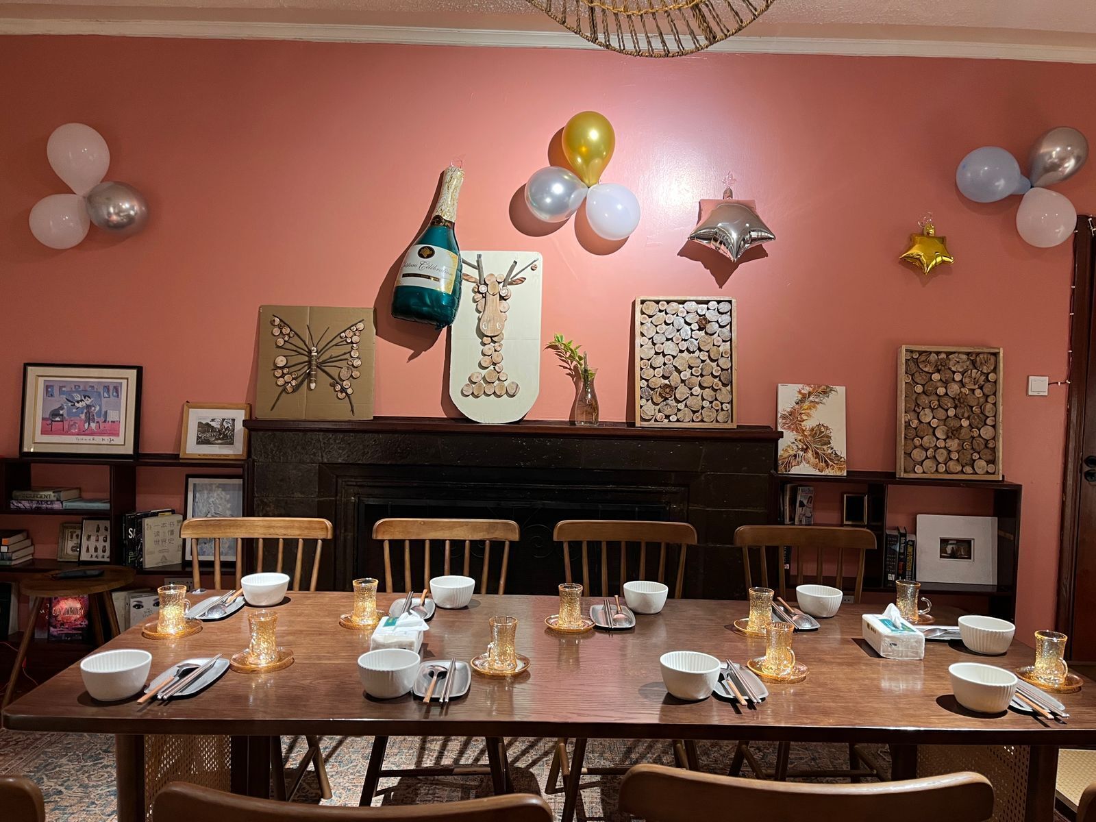 Une salle à manger avec une table en bois dressée pour six personnes, entourée de chaises. On aperçoit une cheminée au fond de la pièce, et les murs sont décorés de divers tableaux encadrés ainsi que d’une grande œuvre d’art en forme de bûche de bois.