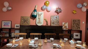Une salle à manger avec une table en bois dressée pour six personnes, entourée de chaises. On aperçoit une cheminée au fond de la pièce, et les murs sont décorés de divers tableaux encadrés ainsi que d’une grande œuvre d’art en forme de bûche de bois.