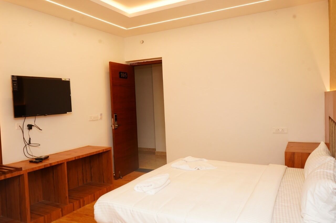 A hotel room with a bed, a TV mounted on the wall, a wooden bench, and a door leading to the hallway.