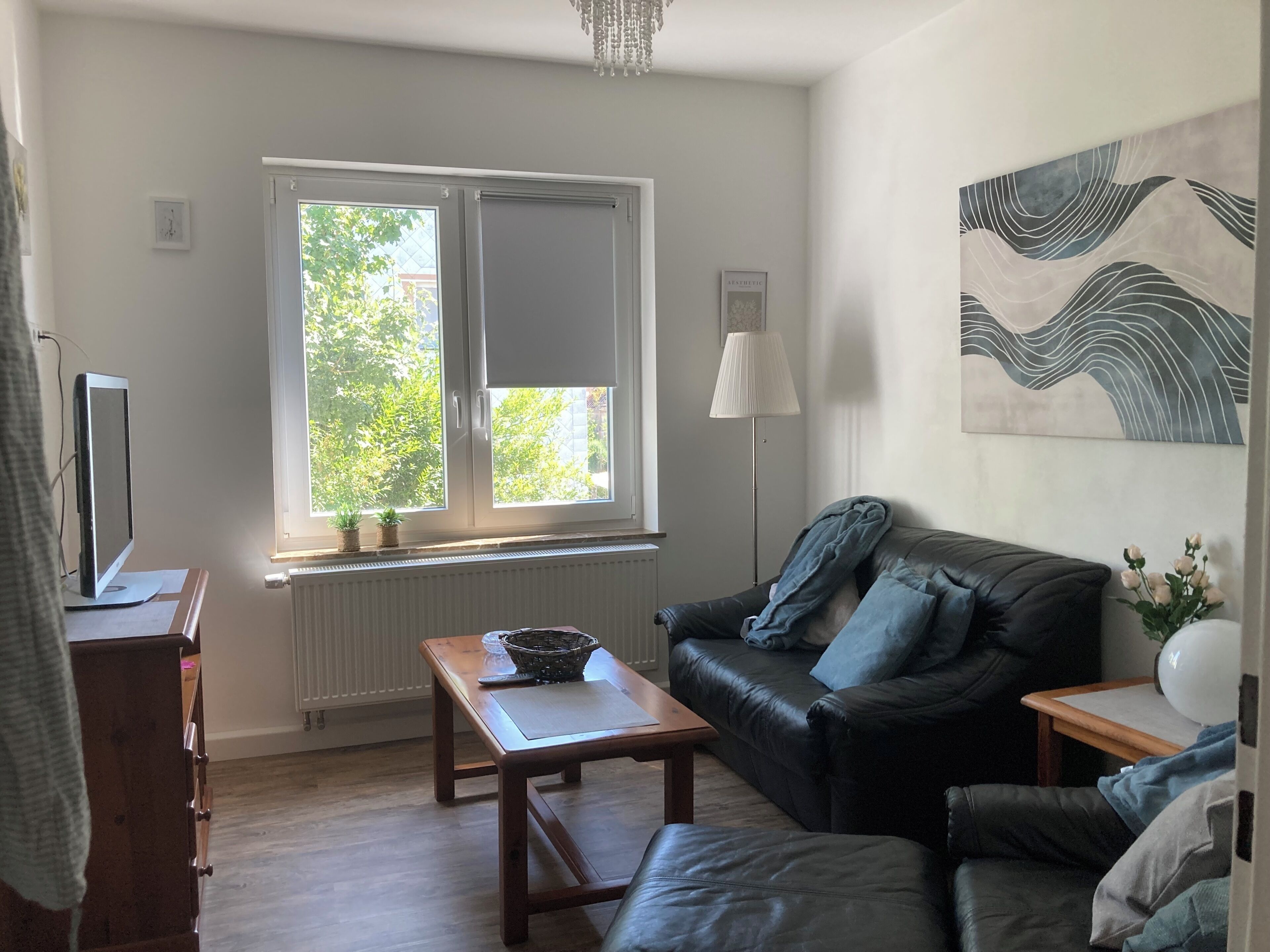 A living room with a black leather sofa, a wooden coffee table, a standing lamp, and a window with a view of greenery.