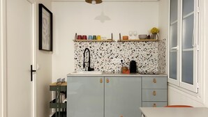 A compact kitchen with a countertop, a sink, and a coffee machine. There is a small table and a chair.