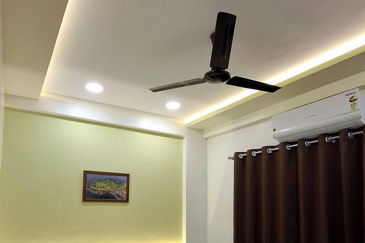 A hotel room with a bed, a ceiling fan, a nightstand, a wall-mounted air conditioner, and a painting on the wall.