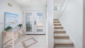 Un couloir avec une porte blanche, un escalier et une console en osier sur laquelle est posée une plante.