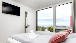 Quarto de hotel com cama, televisão fixada na parede e amplas janelas com vista para o exterior.