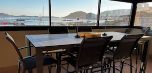 A dining area with a table and chairs, a wooden bowl, and a view of a harbor with boats and mountains.