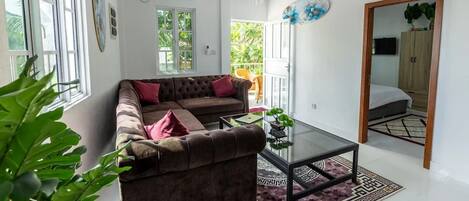 A living room with a brown sofa, a glass coffee table, and a patterned rug. There is a large window with white frames, a ceiling fan, and a door leading to another room.