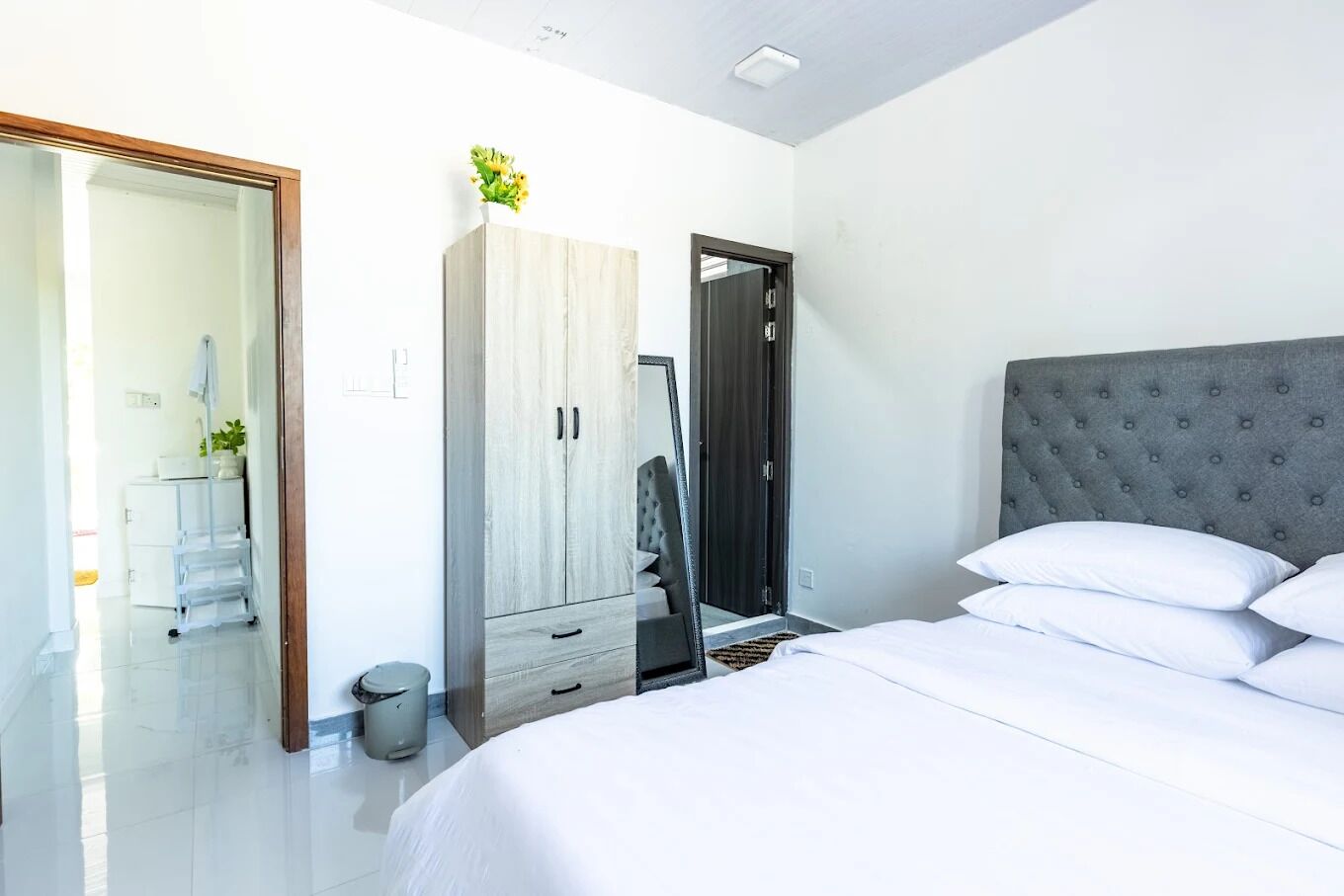 A modern bedroom with a large bed, a grey tufted headboard, a wooden wardrobe, and a mirror.