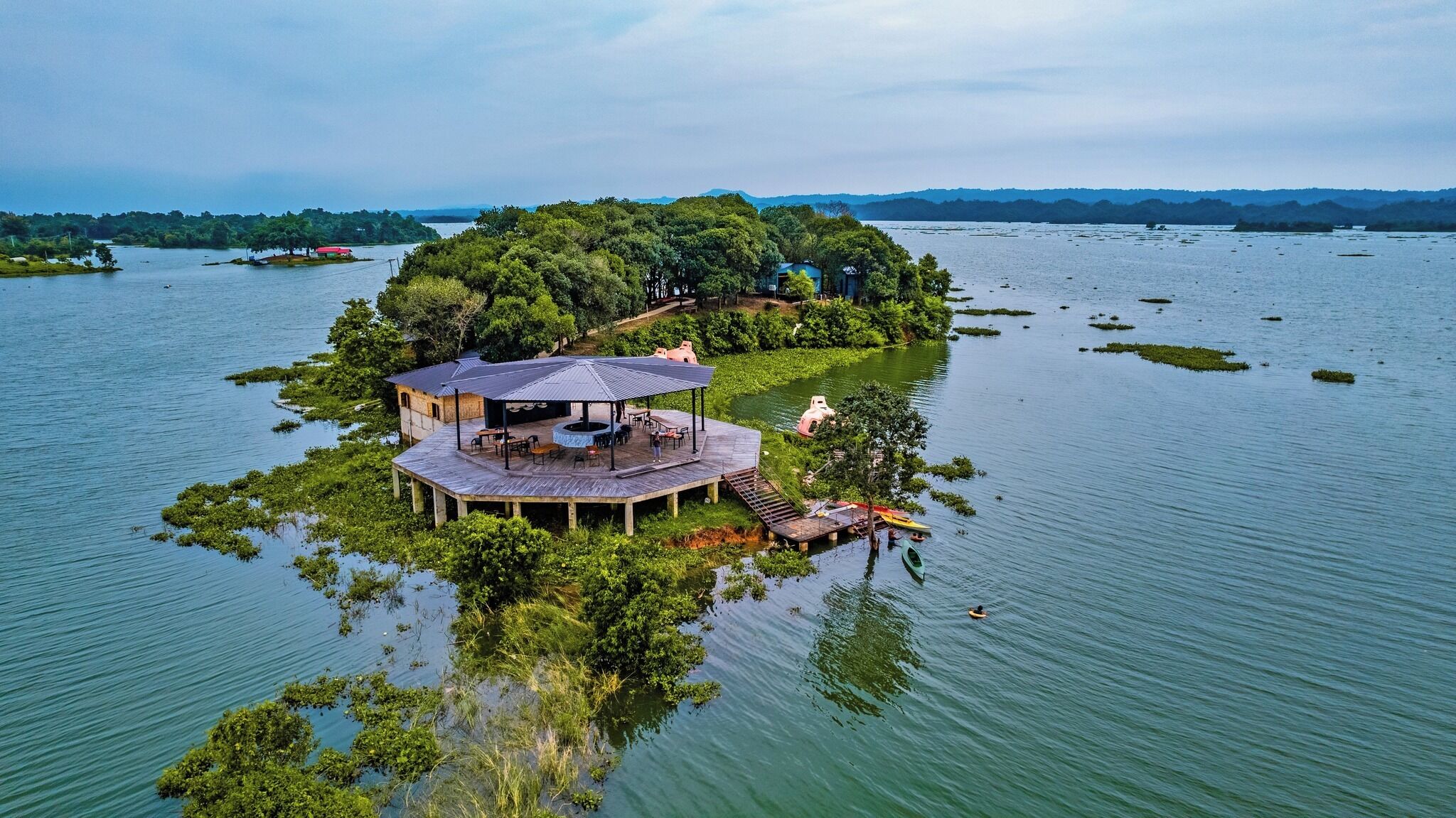 A secluded island with a wooden structure featuring an open deck and seating area, surrounded by water and greenery.