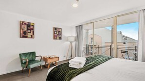 Habitación de hotel con una cama grande, un sillón verde, una mesita de madera con un libro, una lámpara de pie y vistas a la ciudad a través de un ventanal amplio.