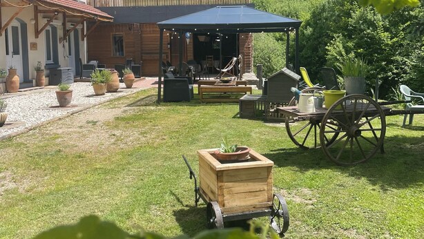 A rustic outdoor seating area with a wooden table and chairs, a covered patio, and a cart with potted plants.