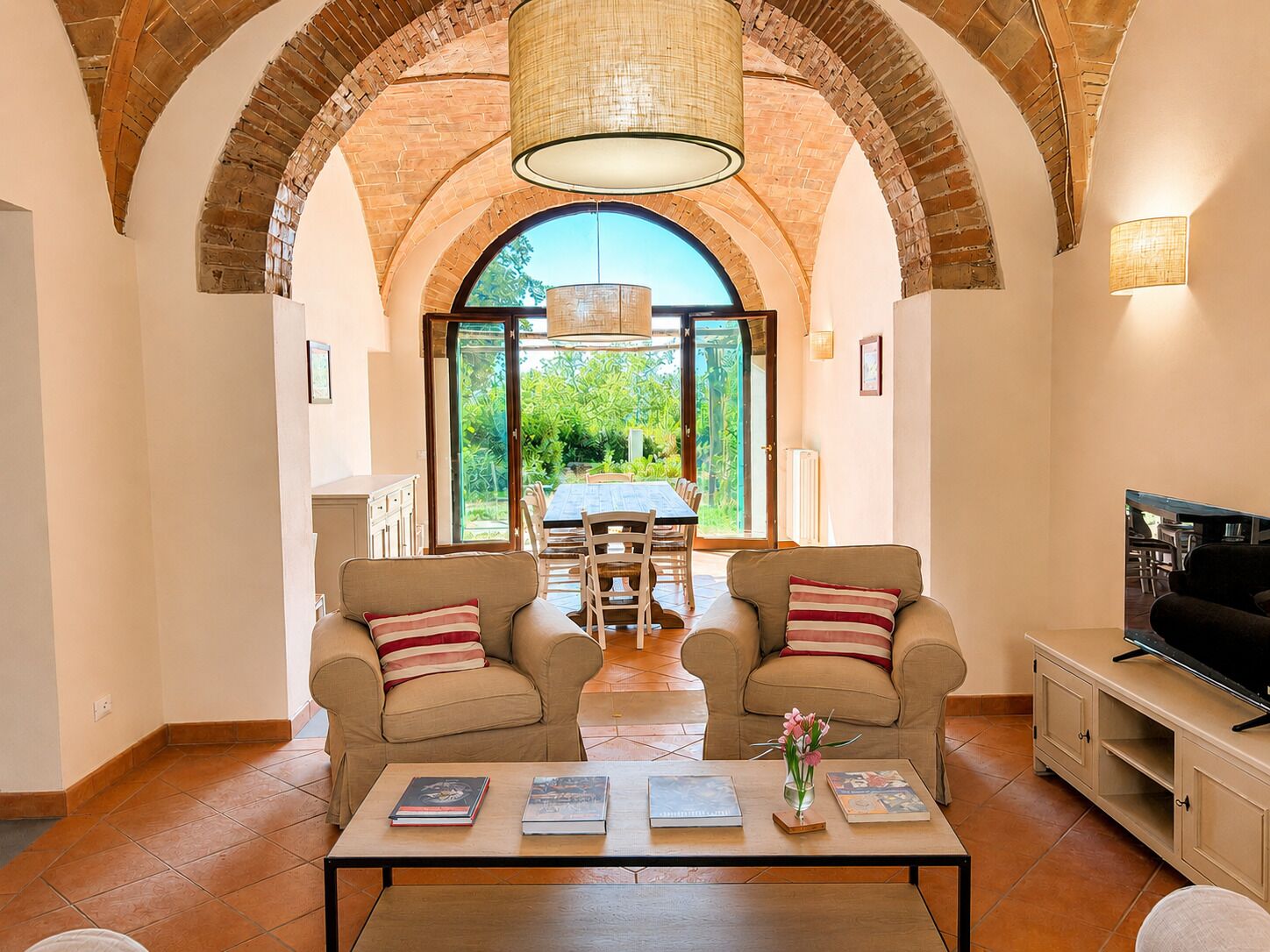 A living room with two armchairs, a coffee table with books and a vase, a TV stand, and a large arched brick ceiling.