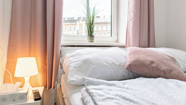 A hotel room with a bed, a bedside lamp, a remote control, a tissue box, and a window with a view of buildings. - Luxury 6BR Apt near Millennium City (Vienna)