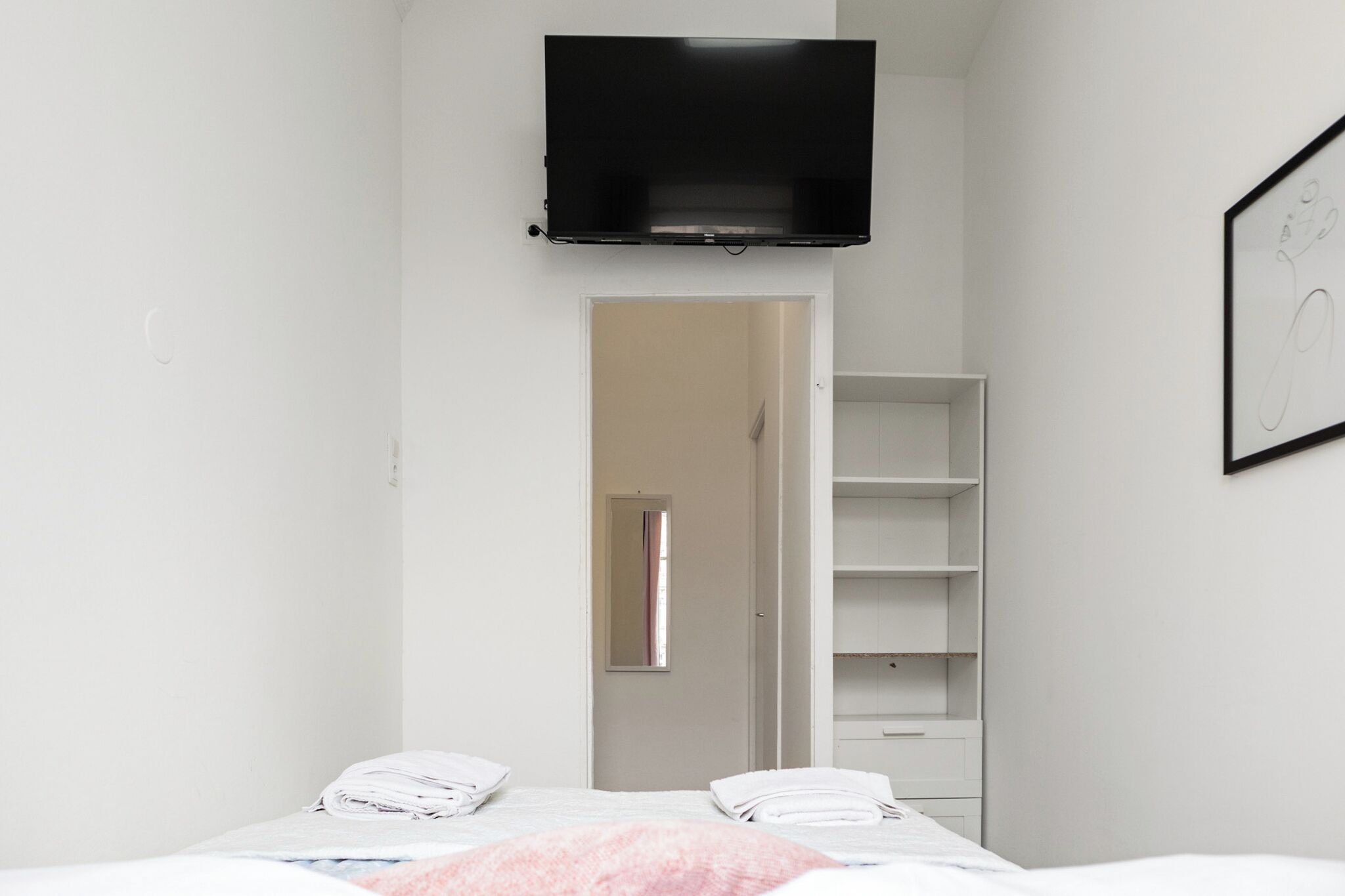 A bedroom with a bed, a TV mounted on the wall, a wardrobe, and a door leading to another room.