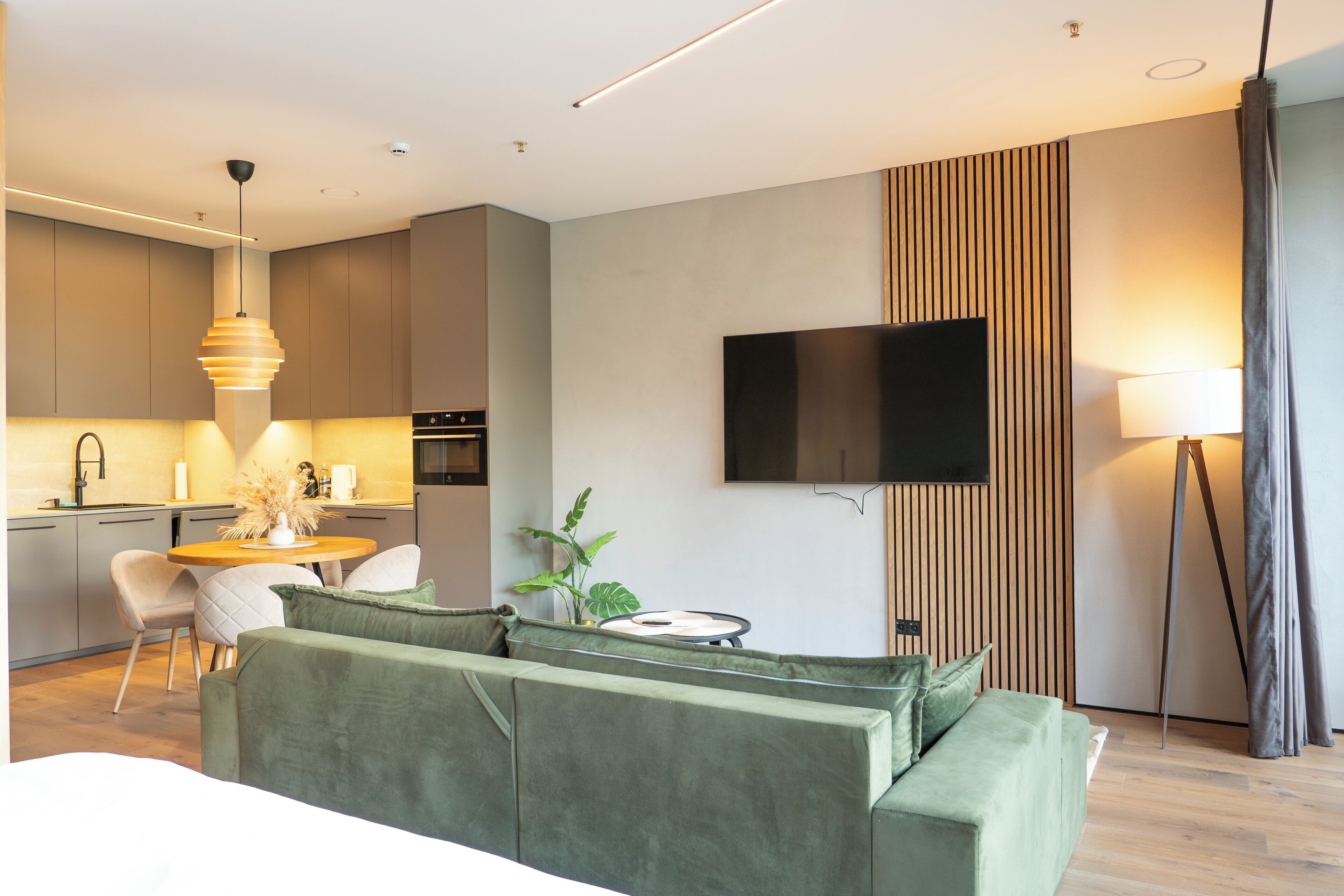 Ein modernes Wohnzimmer mit einem grünen Sofa, einem Esstisch mit Stühlen, einem flachbildfernseher an der Wand und einer Pendelleuchte über dem Tisch.