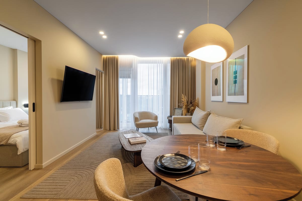 A modern hotel room with a dining area, a sofa, a TV mounted on the wall, and a large window with curtains.