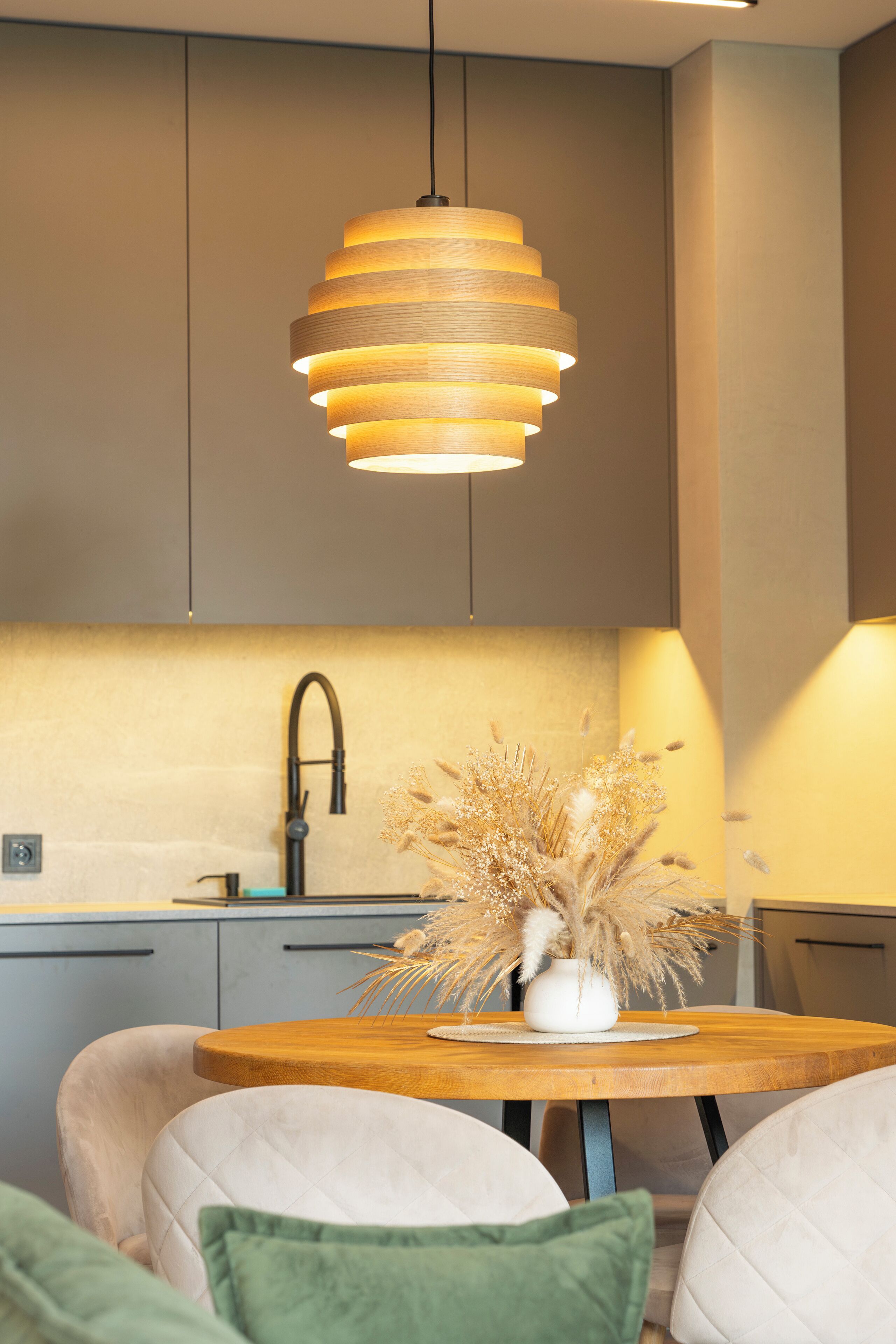 A modern kitchen with a wooden dining table, a vase with dried flowers, and a pendant light.