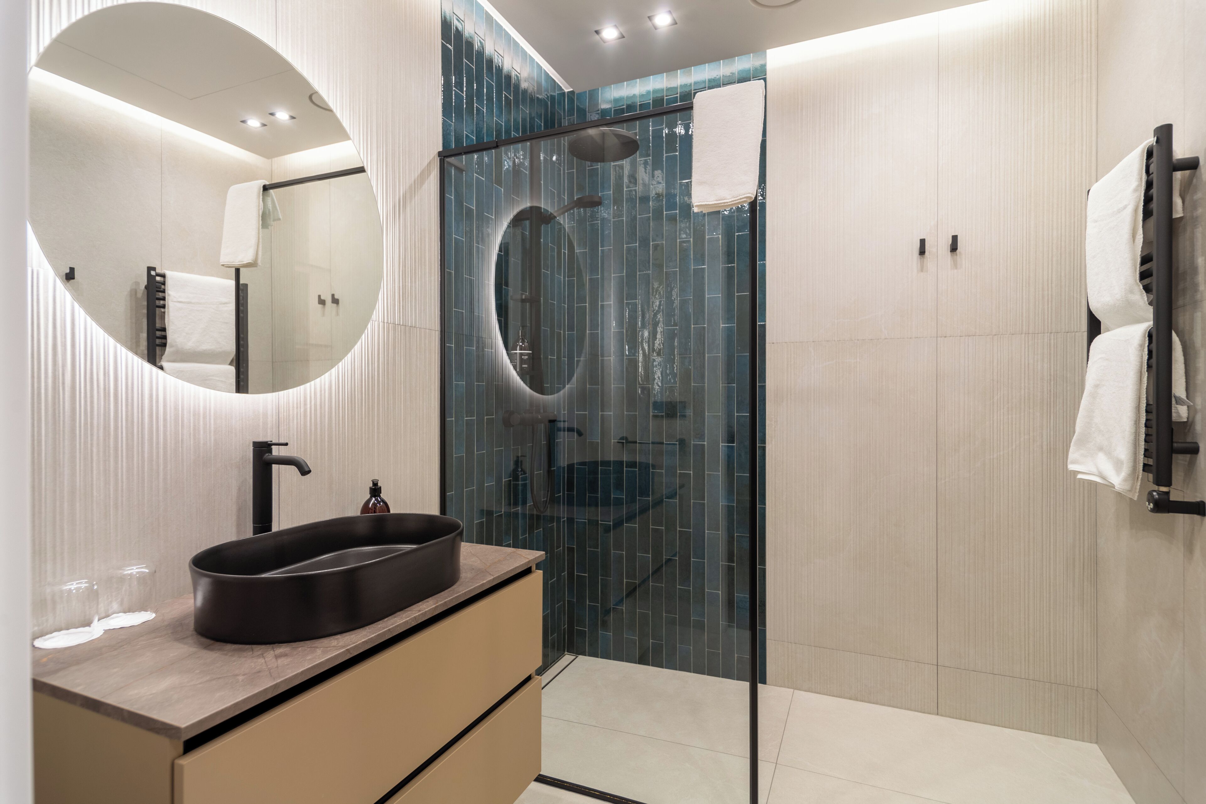 Un baño moderno con un espejo redondo, un lavamanos negro y una ducha con mampara de cristal.