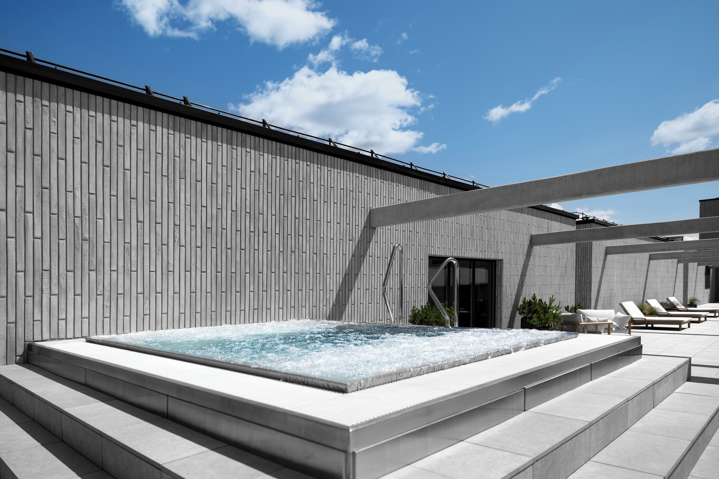 A modern outdoor spa area with a hot tub, lounge chairs, and a concrete wall.